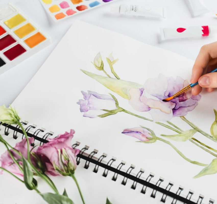 Woman painting eustomas in sketchbook at white table, closeup