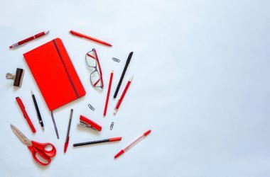 Flat lay of red notebook and stationery on blue background with copy space. Top view of red journal for monthly planning and creativity. School agenda and office supplies, pencils, glasses, scissors.